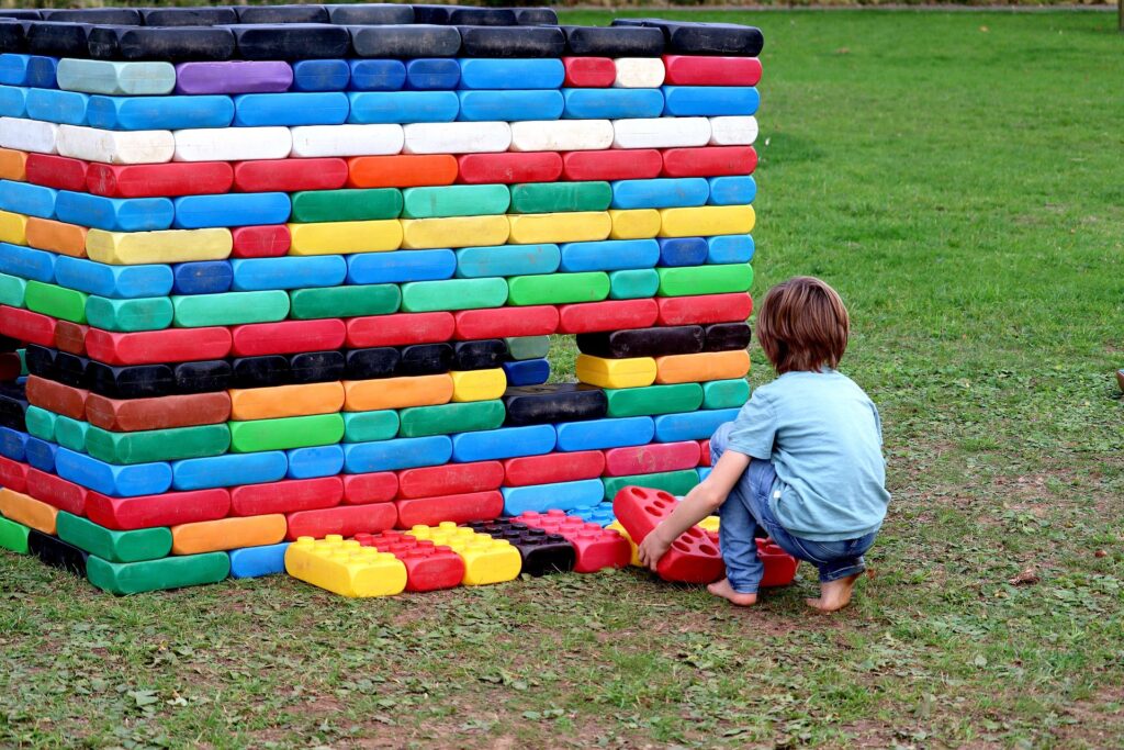 Copil care se joaca in aer liber construind un zid din blocuri mari colorate – joc educativ pentru dezvoltarea motricitatii si gandirii logice