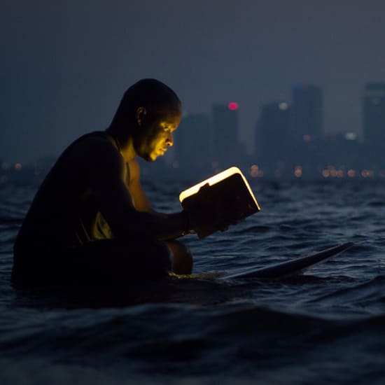 Barbat care citeste o carte luminoasa stand pe o placa de surf in ocean pe timpul noptii, cu profilul orasului luminat in departare